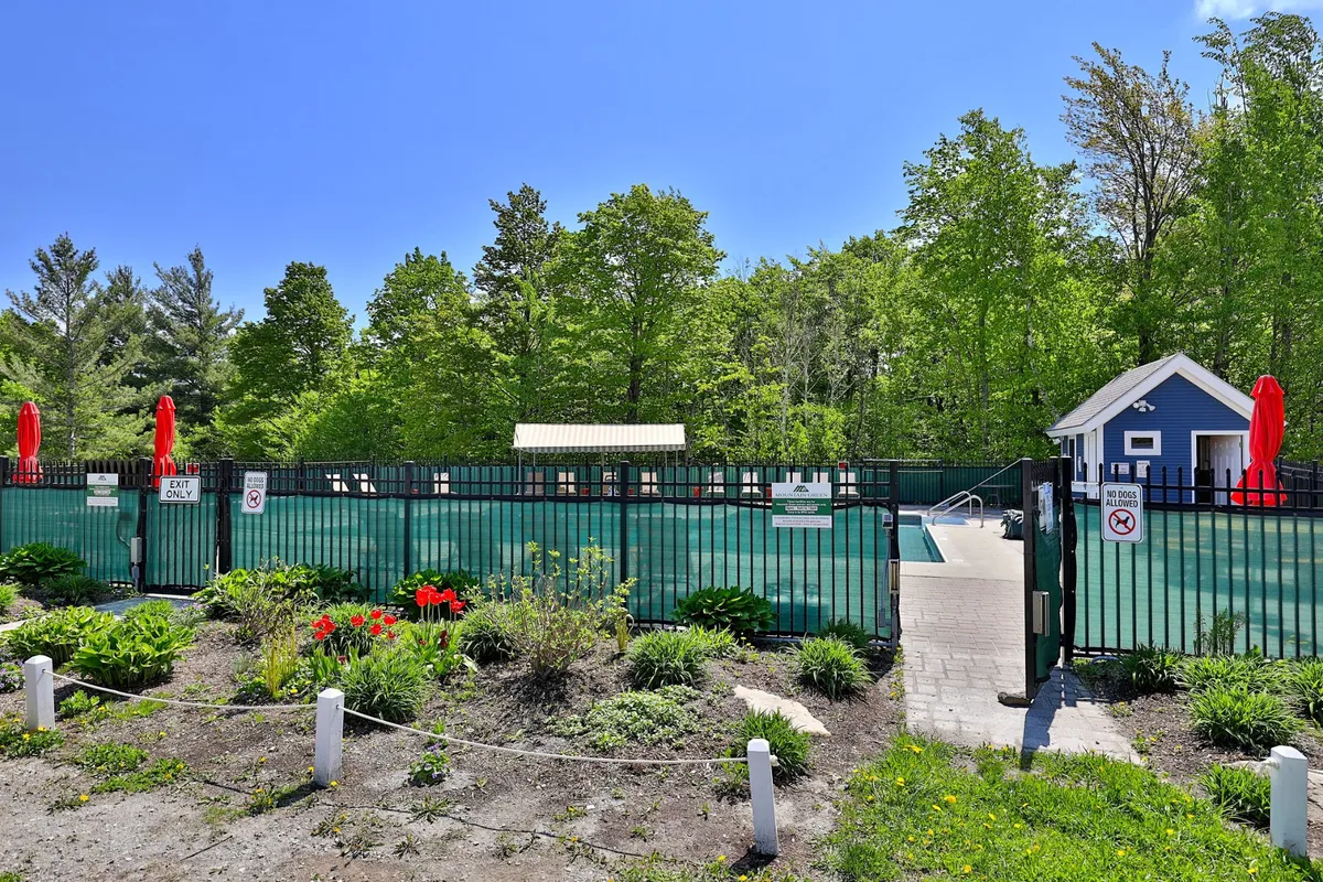 This image captures the vibrant outdoor swimming pool area, surrounded by poolside seating, landscaped gardens, and bright red umbrellas. The fenced area and lush greenery create a lively setting ideal for relaxing or gathering with others during warmer months.