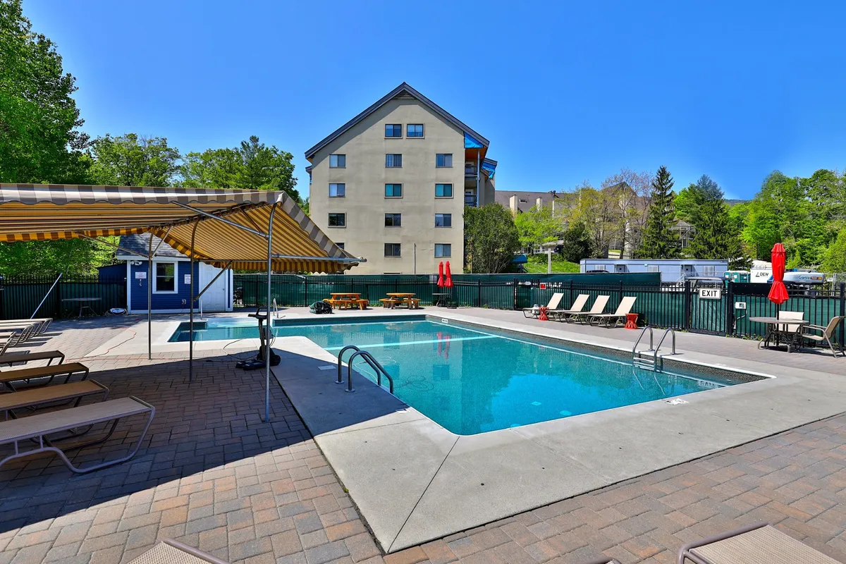 A spacious exterior view featuring a swimming pool with loungers and a cabana, set against a backdrop of green trees and the property building. The scene captures a tranquil, leisurely environment ideal for sunbathing or gathering with friends and family.