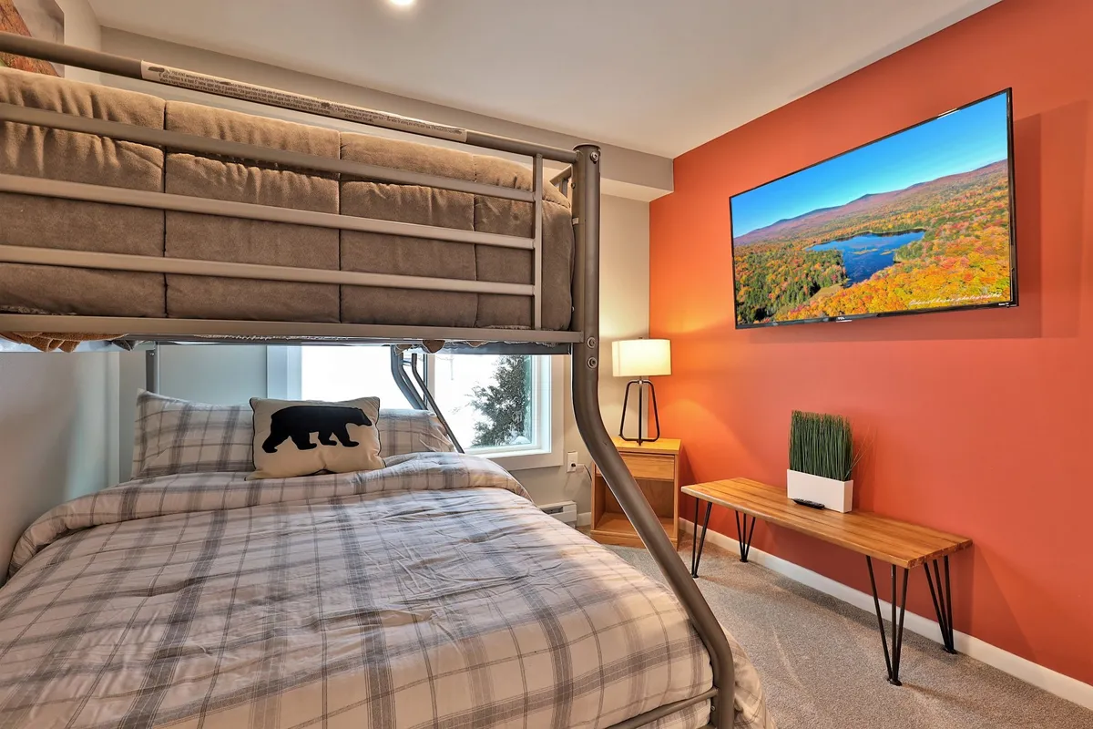 An inviting bunk room with a twin/full bunk bed, a wall-mounted TV, a wooden table with a potted plant, and a window offering scenic views. The space feels perfect for families or groups, with flexible sleeping arrangements and entertainment options.