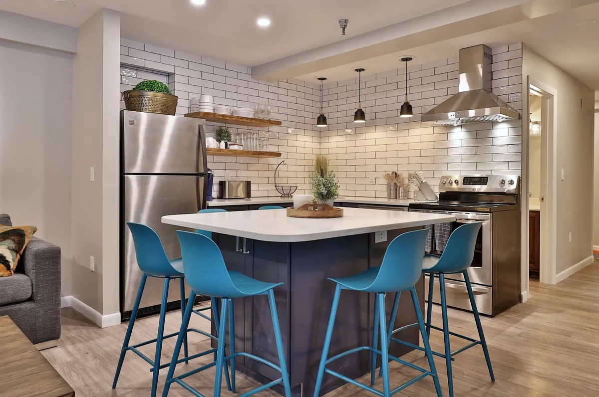 A bright, modern kitchen featuring a central island with blue bar stools, sleek stainless steel appliances, ample counter space, and open shelving adorned with greenery. The scene suggests a welcoming environment perfect for gathering, with sightlines to other living spaces and a hint of mountain ambiance through the windows.