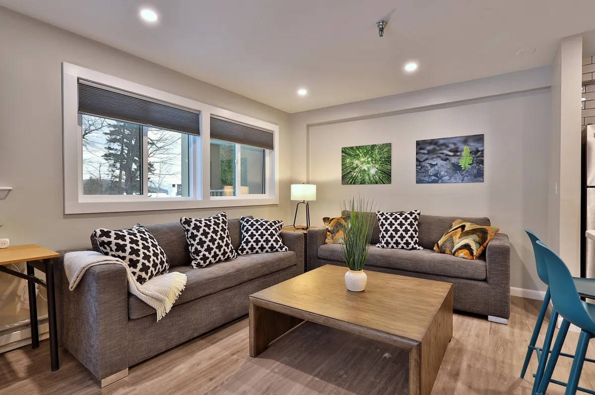 A cozy living room with a plush sofa, a coffee table, art on the walls, a floor lamp, and windows letting in natural light. The setting invites guests to relax, socialize, or convert the sofa into an extra sleeping space if needed.