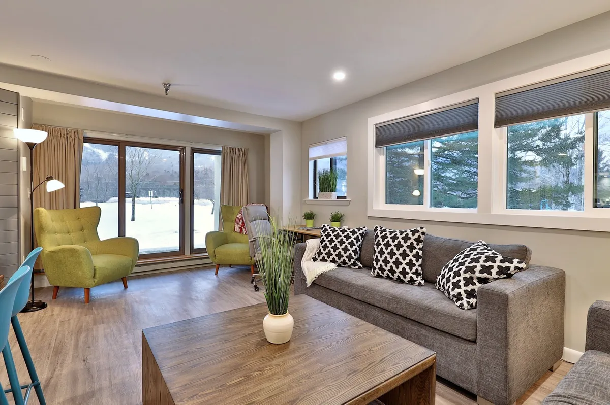 The image displays a bright, cozy living area furnished with green armchairs, a decorative sofa with cushions, and a wooden coffee table. Large windows flood the room with natural light and provide stunning mountain views, reinforcing the connection to the outdoors. The decor evokes comfort and togetherness.