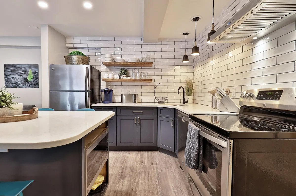 A modern, inviting kitchen featuring a spacious island countertop, a suite of stainless steel appliances, and shelves with decorative items. The layout highlights ample work and gathering space, ideal for both everyday cooking and special occasions.