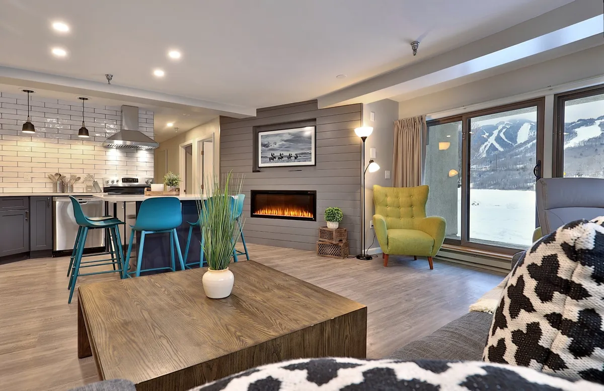 The image showcases a modern, renovated kitchen with a central island, a cozy fireplace, colorful accent chairs, and expansive windows revealing stunning mountain views. The scene exudes warmth and comfort, with plenty of natural light and a clear view of the sunrise over snow-capped peaks, inviting guests to relax and gather with friends or family.