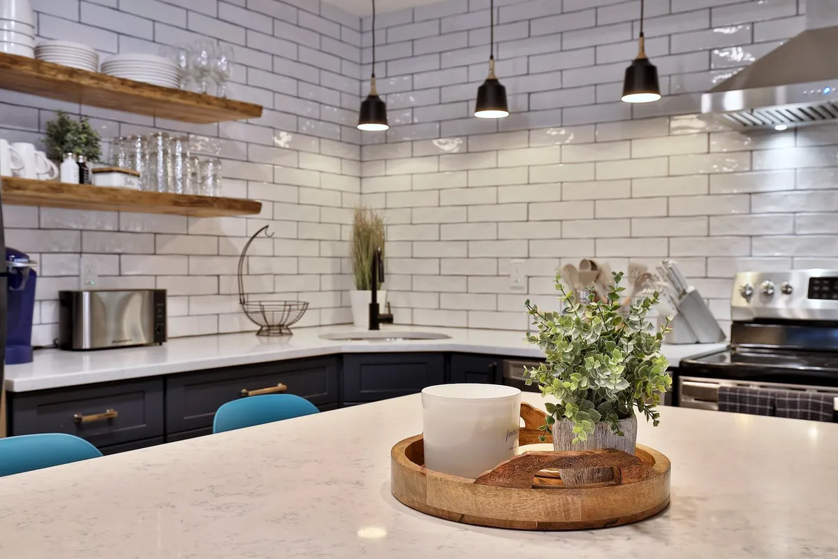 A contemporary kitchen scene with a central island topped by a decorative tray and small plants, shelves filled with dishes and glasses, and gleaming stainless steel appliances along the back wall. The room exudes style and functionality, capturing the essence of a kitchen ready for group gatherings and meal preparation.