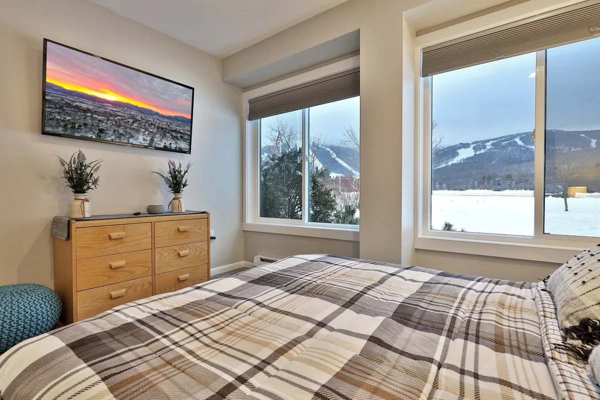 A serene queen bedroom featuring a comfortable bed with plaid bedding, large windows looking out to scenic mountain views, a mounted TV displaying a sunset image, and a chest of drawers for storage. The ambiance feels tranquil and perfect for unwinding after an active day.